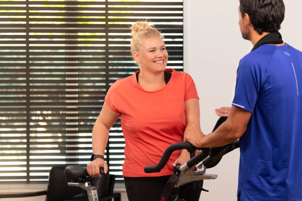 A smiling woman in an orange shirt interacts with a male instructor in a blue shirt while resting her hands on a stationary bike in a gym setting.
