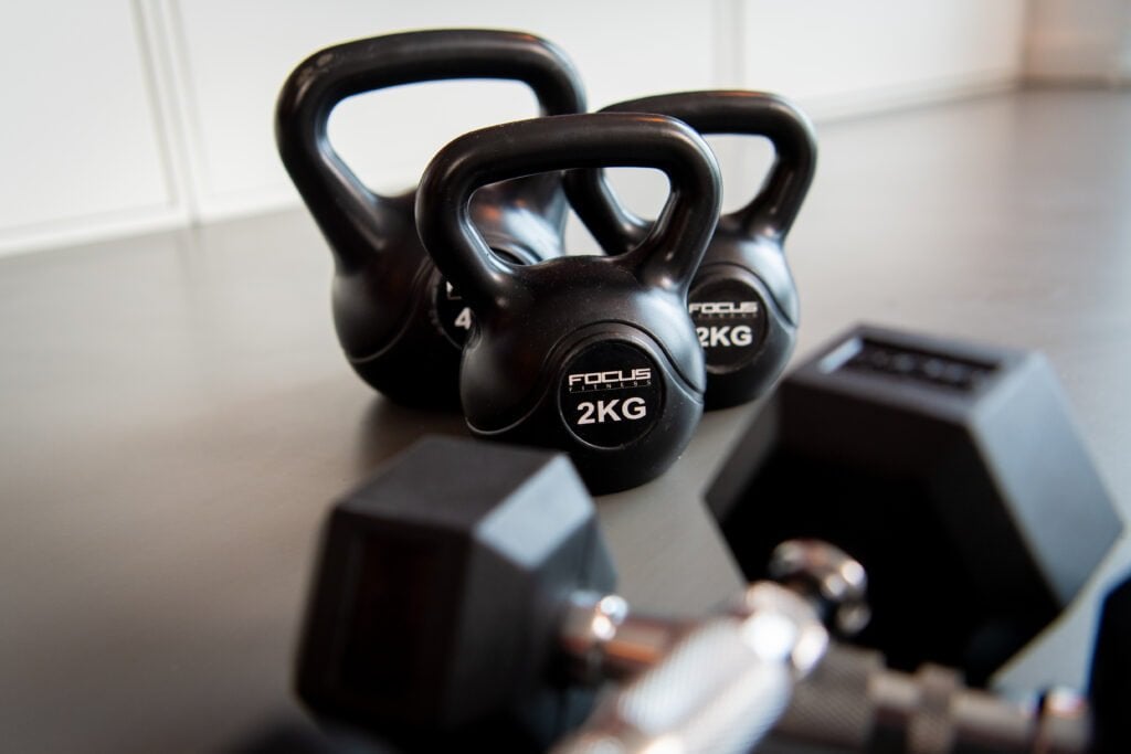 Two black kettlebells labeled "2 kg" on a gym floor, with a dumbbell in the foreground, emphasizing a focus on fitness equipment.