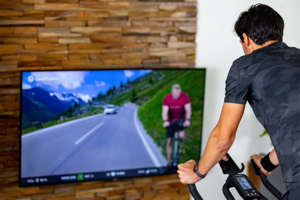 A man in a black t-shirt is cycling on a stationary bike indoors, facing a large screen displaying a virtual cycling route with scenic views.