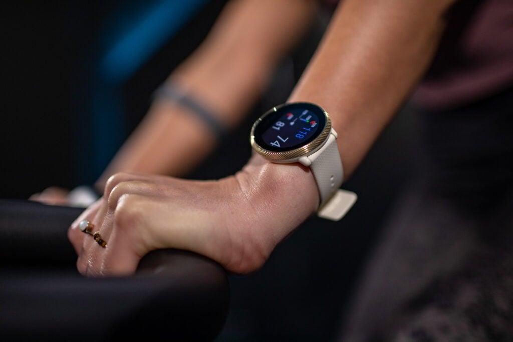 Close-up of a smartwatch on a woman's wrist, displaying time and heart rate, while she grips a handlebar, indicating an exercise setting.