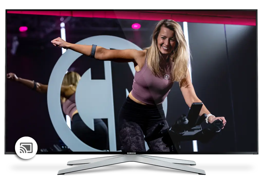 A woman with blond hair, wearing a purple top and black pants, is energetically indoor cycling in front of a large lit 'g' logo, seen on a laptop screen.
